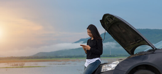 Asian woman with a broken car with open hood.Young woman using mobile phone while looking at broken down car on street