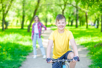 Obraz premium Happy boy rides on a bicycle in summer sunny park. Mom stands behind on the background