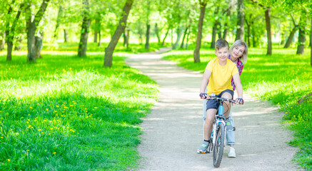 Happy family. Sporty family leisure. Mom teaches her young son ride a bike in summer park. Panoramic view. Empty space for text
