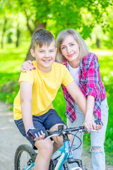 Fototapeta premium Sporty family leisure. Happy mother embraces her young son, who ride bikes in a summer park