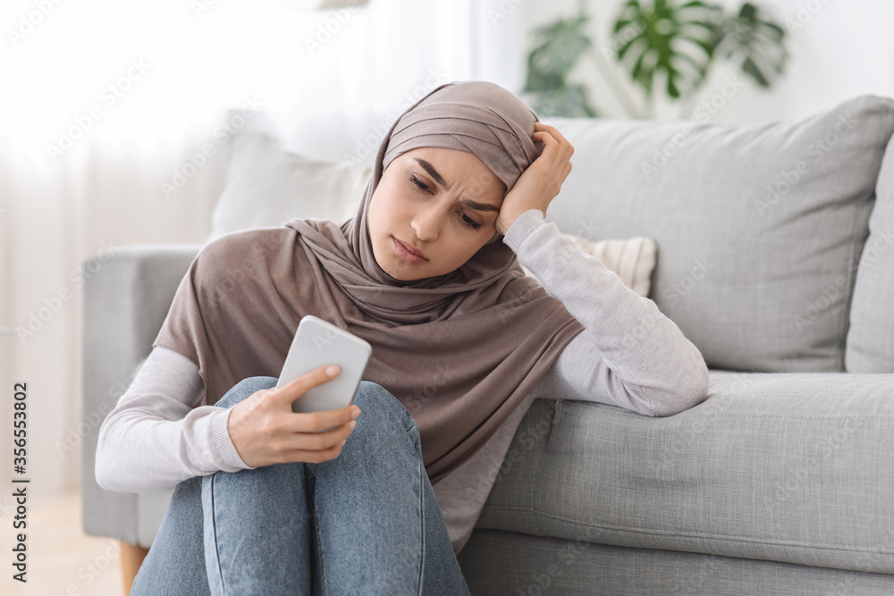 Wall mural Depression Concept. Upset Arabic Woman Sitting With Smartphone On Floor At Home