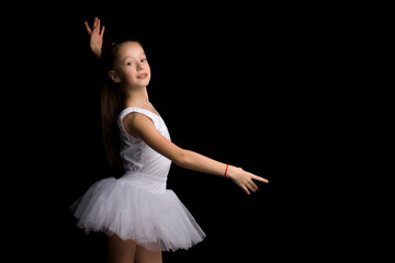 Fototapeta premium Cute little girl in a tutu and pointe shoes is dancing in the studio on a black background.