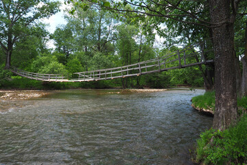 A wooden suspension bridge over a small picturesque river flowing in a wooded area. Summer landscape.