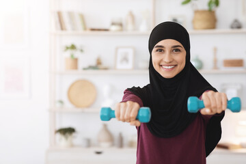 Smiling muslim girl in hijab exercising with dumbbells at home