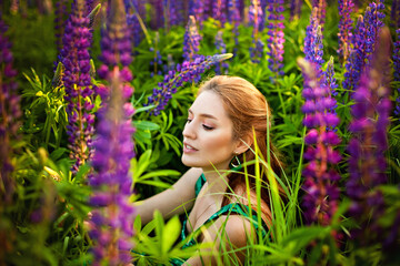 A young beautiful girl with red hair is sitting with a purple Lupin in a blooming field. Blooming Lupin flowers.