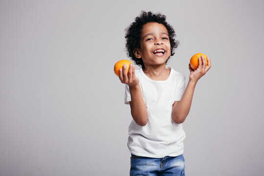 Laughing dark-skinned Moroccan boy holds in his hands two juicy oranges on a light background