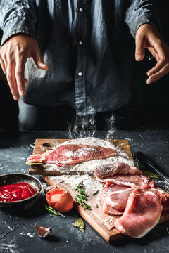 Raw chops and pieces of pork for escalopes on cutting boards with flour, rosemary, bay leaf, sauce on the kitchen table. One piece of meat fell from the chef's hands into flour. Place for text