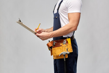 repair, construction and building - male worker or builder with clipboard, pencil and working tools on belt over grey background