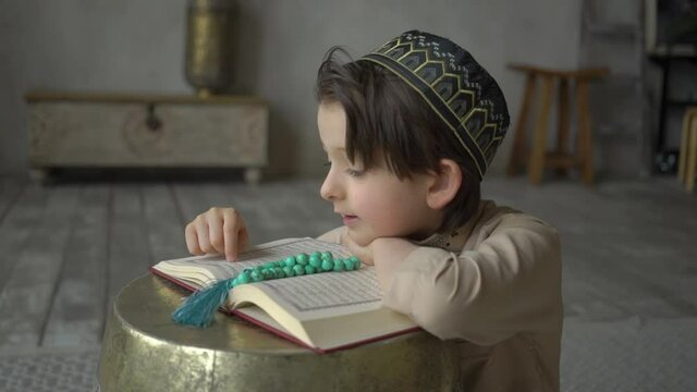 Little boy reading the Koran in prayer hat and arabic clothes with rosary beads and Quran book praying to Allah, prophet Muhammad holy spirit religion symbol concept inside eastern interior