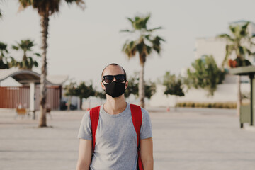 Young male traveler with backpack and in sun glasses wearing black protective mask. Covid 19 or coronavirus pandemic concept. Man in a dust protection reusable  mask