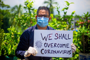 A young man wearing a medical mask and safety gloves stands with a placard message to "We shall overcome Coronavirus".A man holding a placard message in outdoor natural light.