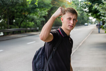 Portrait of a young man in a black T-shirt looking to camera with raising his hand with the clenched fist on the background of a road and green trees.