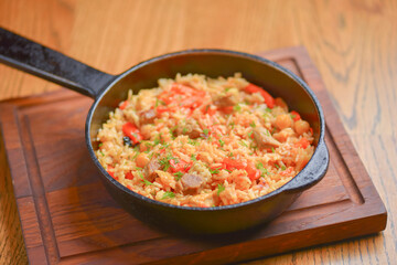Rice with meat and vegetables served in a frying pan on wooden board over light rustic wooden table