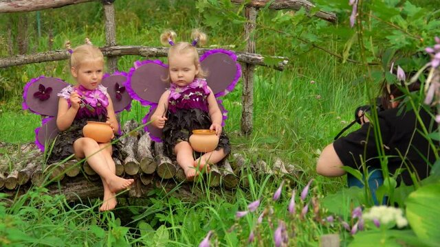 Female photographer takes pictures of cute girls playing purple butterflies and eating honey. Girls smile and wear handmade butterfly wings