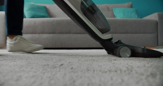 Close up of female legs in jeans and white sneakers using vacuum cleaner for washing white carpet at home. Housewife cleaning home from dust with modern device.