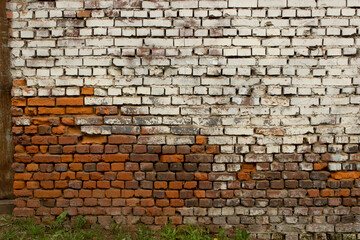 Old damaged brick wall in two colors, half white, the second half red. Uneven sloppy brickwork. A little green grass below.