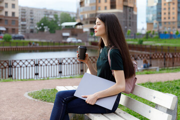 Relaxed young woman in a green T-shirt and with a backpack sitting on a wooden bench, drinking coffee and browsing on her laptop