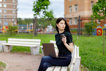 Fototapeta premium Relaxed young woman in a green T-shirt and with a backpack sitting on a wooden bench, drinking coffee and browsing on her laptop