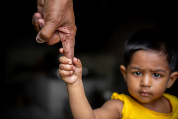 A baby boy confidently walks around holding on the finger of a senior old man. Family, Generation, Support and people concept. Dark background.