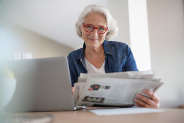 Senior woman reading newspaper in front of laptop