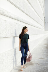 Obraz premium Brunette girl in jeans, a green T-shirt, a hair band and a pink backpack stands against a white wall