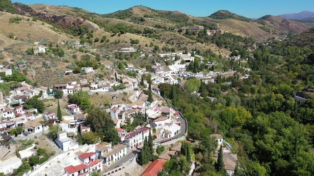 Aerial shot of street by houses and trees in city by mountains against sky, drone flying forward over residential buildings on sunny day - Granada, Spain