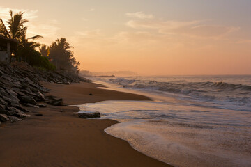 Beautiful postcard landscape shot of the Pitiwella beach (Sri Lanka) at sunrise with orange sand, green palm trees, brown rocks, the Indian ocean with big white waves and the sun on the horizon