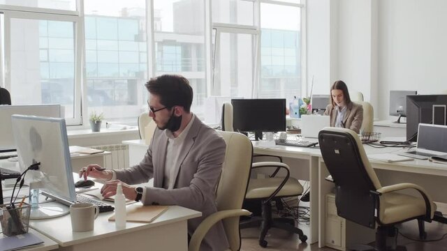 Medium shot of businessman and businesswoman in formalwear and face masks pushed to their chins sitting at desks in empty office and working during covid-19 pandemic