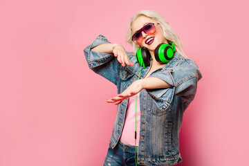 Happy stylish blond woman in pink glasses with big headphones listening music and dancing on colored background
