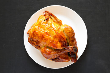 Homemade traditional rotisserie chicken on a white plate on a black background, top view. Flat lay, overhead, from above.