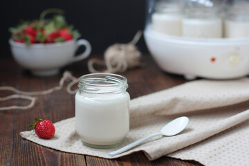 A glass jar of home made yoghurt decorated with no decoration on natural color fabric. One strawberry nearby, more strawberries in blur, a yoghurt maker in blur.