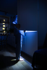 Night hunger and insomnia concept. A girl waking up at night is standing by the refrigerator in the...