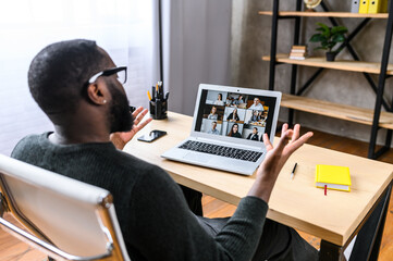 Virtual meeting with many people together. African-American young guy talking online with employees via video connection. Multiracial team. Back view