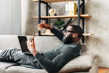 Serious African-American guy in glasses with airpods earphones lays on the sofa in stylish living room and using trendy laptop for video call. Remote work concept