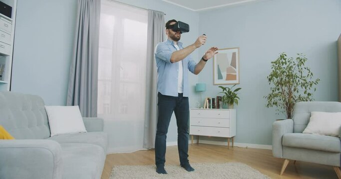 Handsome beautiful man wearing virtual reality headset and holding controllers scrolling virtual display at home. Man staying in VR googles in the middle of living room.