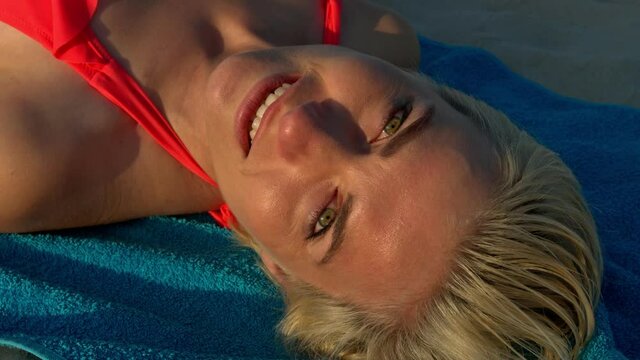 A beautiful young woman with short blond hair, lies on a towel, wearing a bikini on a sandy beach and looks at the camera with a smile during sunset.