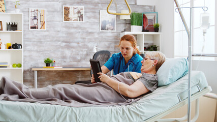Female caregiver helps an old disabled woman lying in hospital bed to use a digital tablet PC....