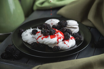 Still life, marshmallows and meringues on a black plate decorated with red berries and syrup. On a dark wooden table. The folds of the fabric on the dark boards.