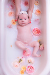 Fototapeta premium Happy kid girl takes a milk bath with petals. Bouquets of pastel rose flowers. Infant bathing. Hygiene and care for young children.
