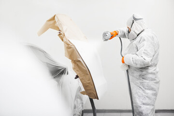 Auto mechanic worker painting a white car with spray gun in a paint chamber during repair work.