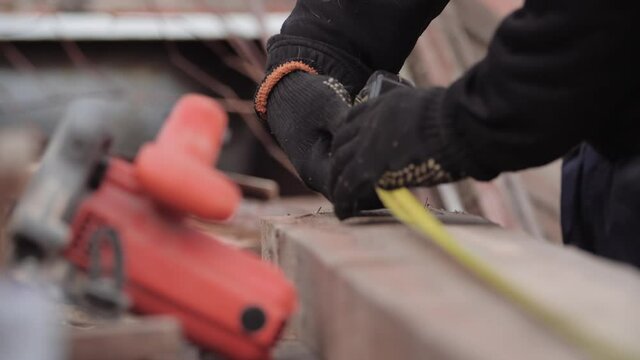 Close-up of unidentified worker in work gloves measuring the length and width of board with tape measure for processing and sawing with a circular. Frame housing construction. Repair with a grinder.
