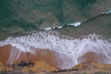 Fototapeta na wymiar aerial drone bird view shot of the sea shore with yellow sand, black rocks, large white waves and foam crashing on the beach forming beautiful textures, patterns, shapes. Sri Lanka