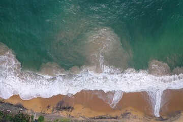 Naklejka na ściany i meble aerial drone bird view shot of the sea shore with turquoise blue water, large white waves and foam, empty beach with yellow sand, black rocks forming beautiful textures, patterns, shapes. Sri Lanka