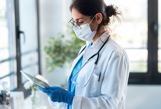 Beautiful Female Doctor Wearing A Hygienic Face Mask And Protective Gloves While Using Her Digital Tablet In The Consultation.