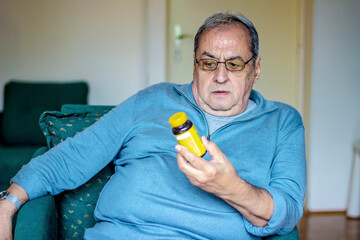 Senior man sitting on sofa at home taking medication. Pensioner reads the description of a medicine...
