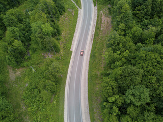 Drone point of view: aerial view flying over two lane countryside forest road with orange car moving green trees of dense woods growing both sides.