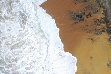 aerial drone bird view shot of the sea shore with yellow sand, black rocks, large white waves and foam crashing on the beach forming beautiful textures, patterns, shapes. Sri Lanka