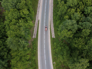 Drone point of view: aerial view flying over two lane countryside forest road with orange car moving green trees of dense woods growing both sides.