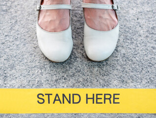 Top view of women feet in white leather shoes stands at yellow sign with text stand here to keep a distance between people.