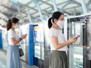 Two Asian women wearing medical face mask, using smartphone waiting for metro at train station platform, standing distance apart from other people.
